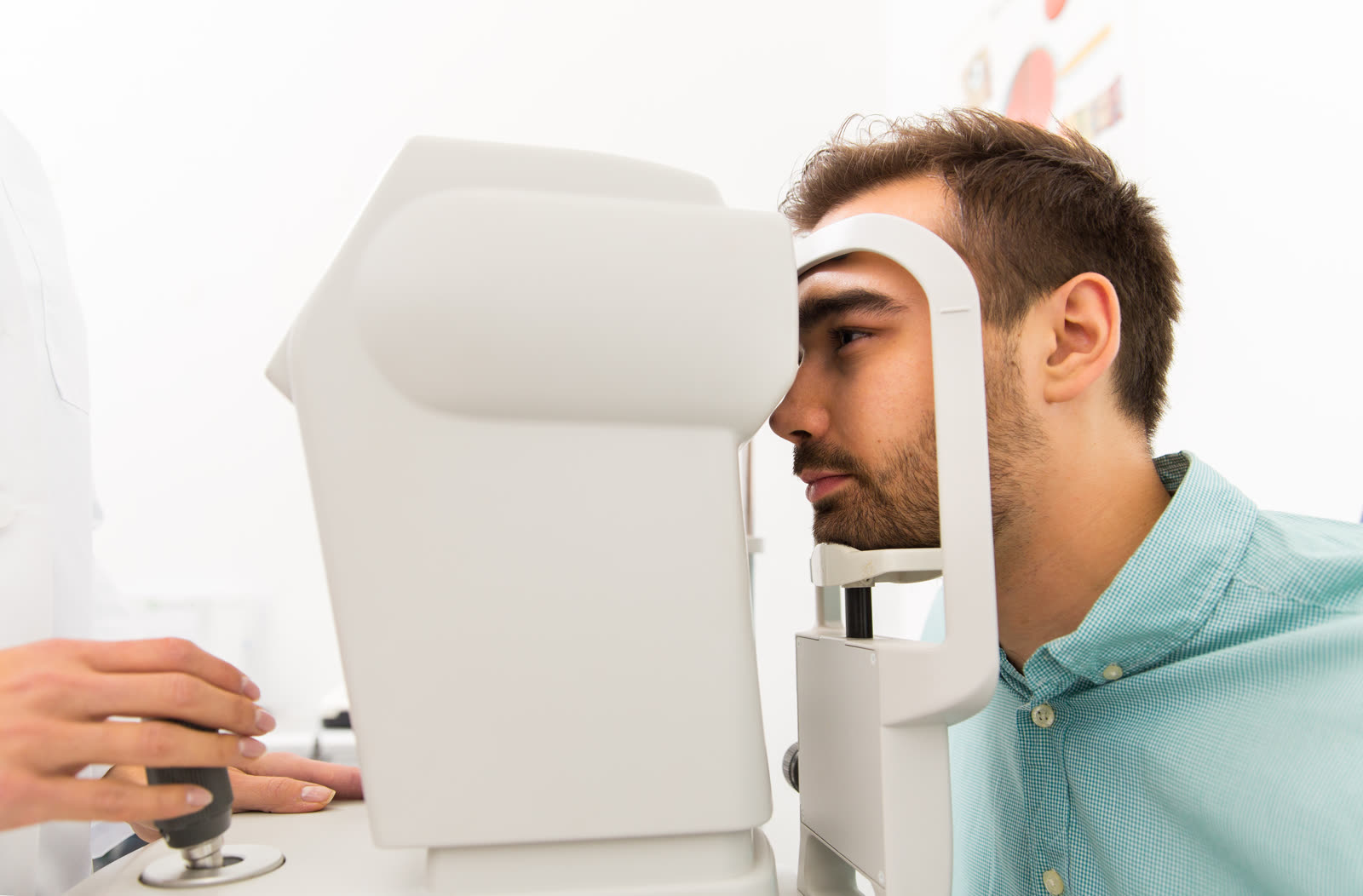 A man leans into an eye-exam instrument with his forehead against the headrest while a technician's hand adjusts the control in a bright exam room.