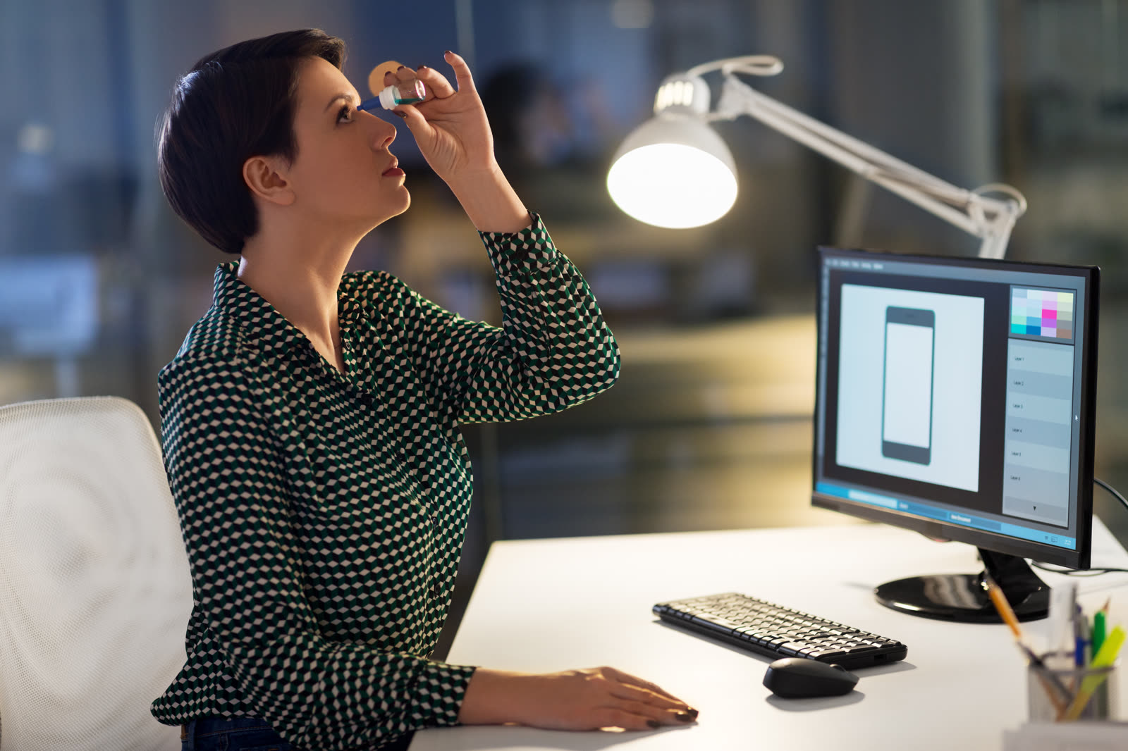 A woman at an office desk tilts her head back and squeezes a drop from a small bottle into her eye, lit by a desk lamp beside her monitor.